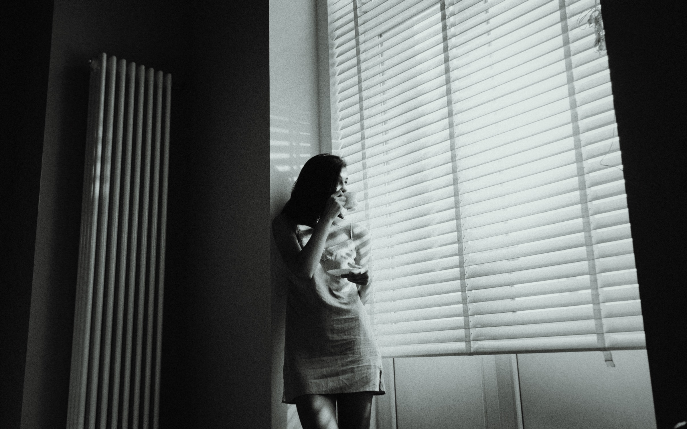 A woman holding a cup by a window with half-open blinds, in soft side light