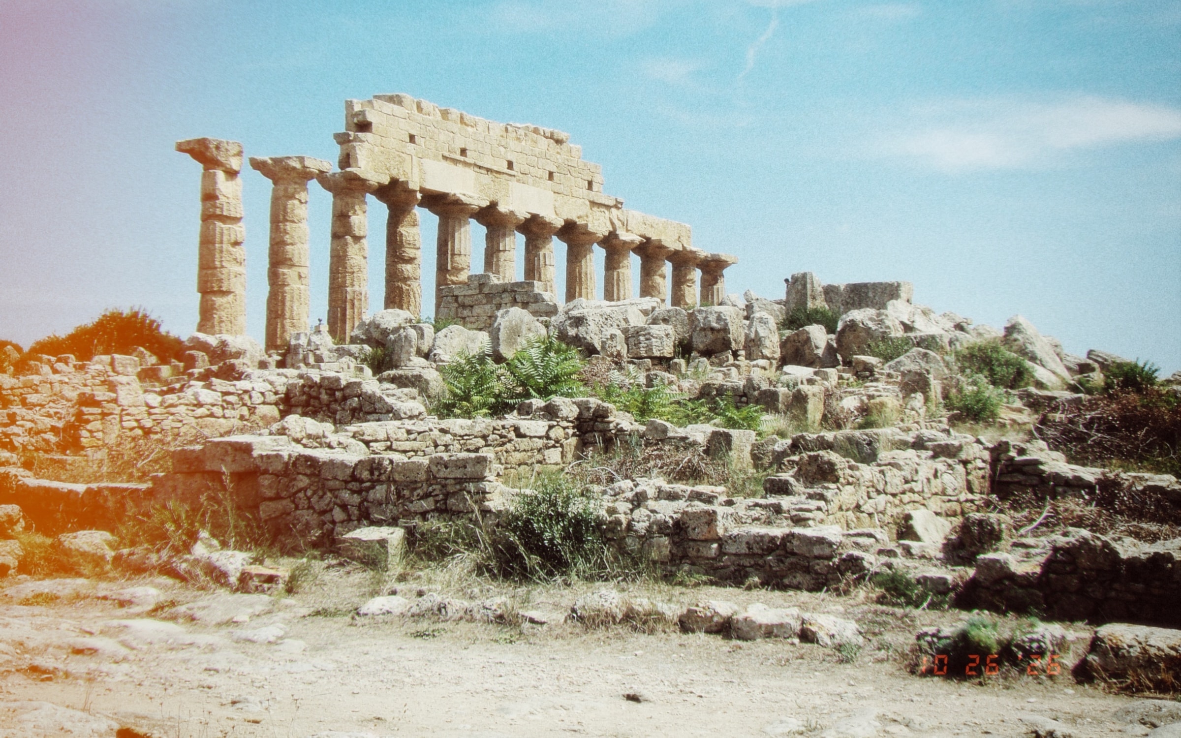The columns of an ancient Greek temple at Selinunte, Sicily, under a blue summer sky, with a warm light leak at the left edge and the date in the corner