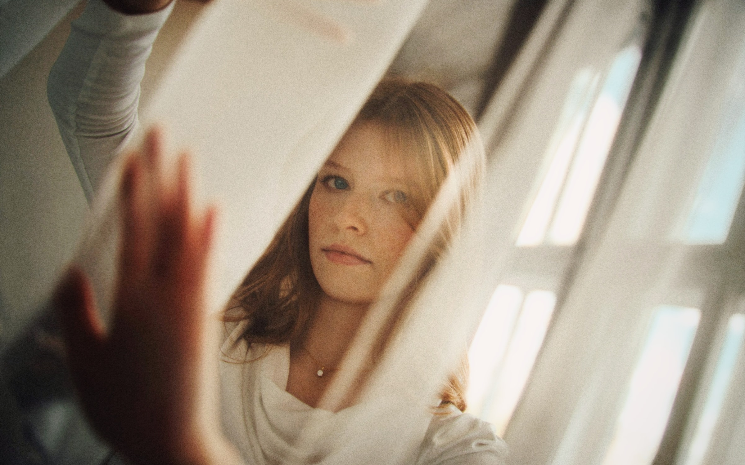 A young woman looking through a sheer curtain in soft window light, her hand on the fabric
