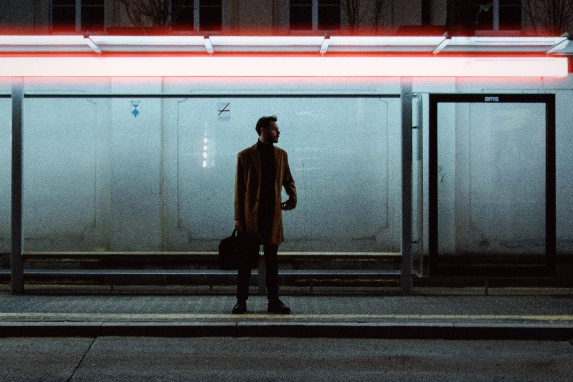 A man in a long coat waiting under the strip lights of a bus shelter at night, rendered with the Nightcall preset