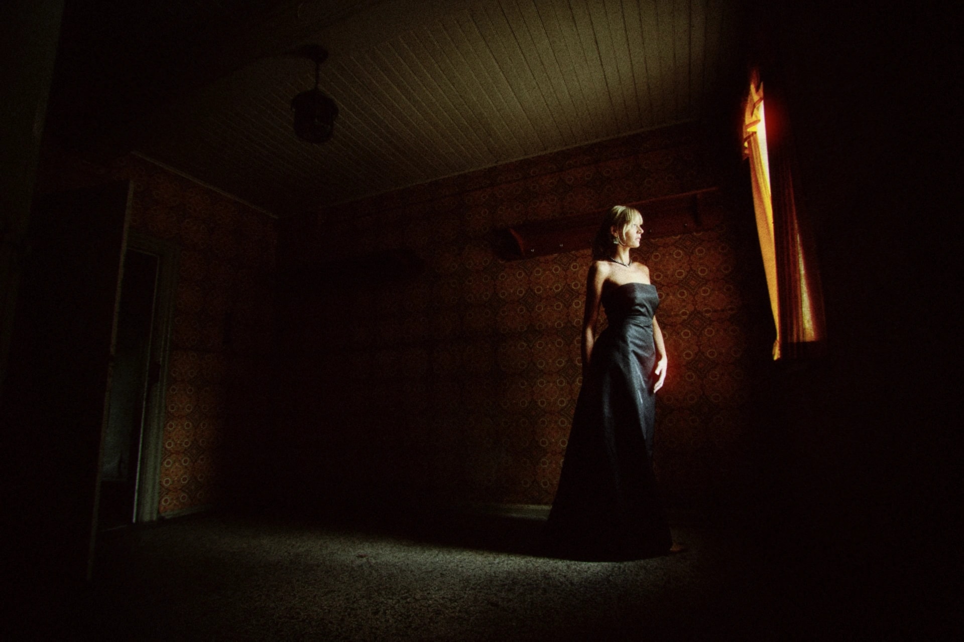A woman in a dark gown standing beside a narrow window of warm light, rendered with the Nightcall preset