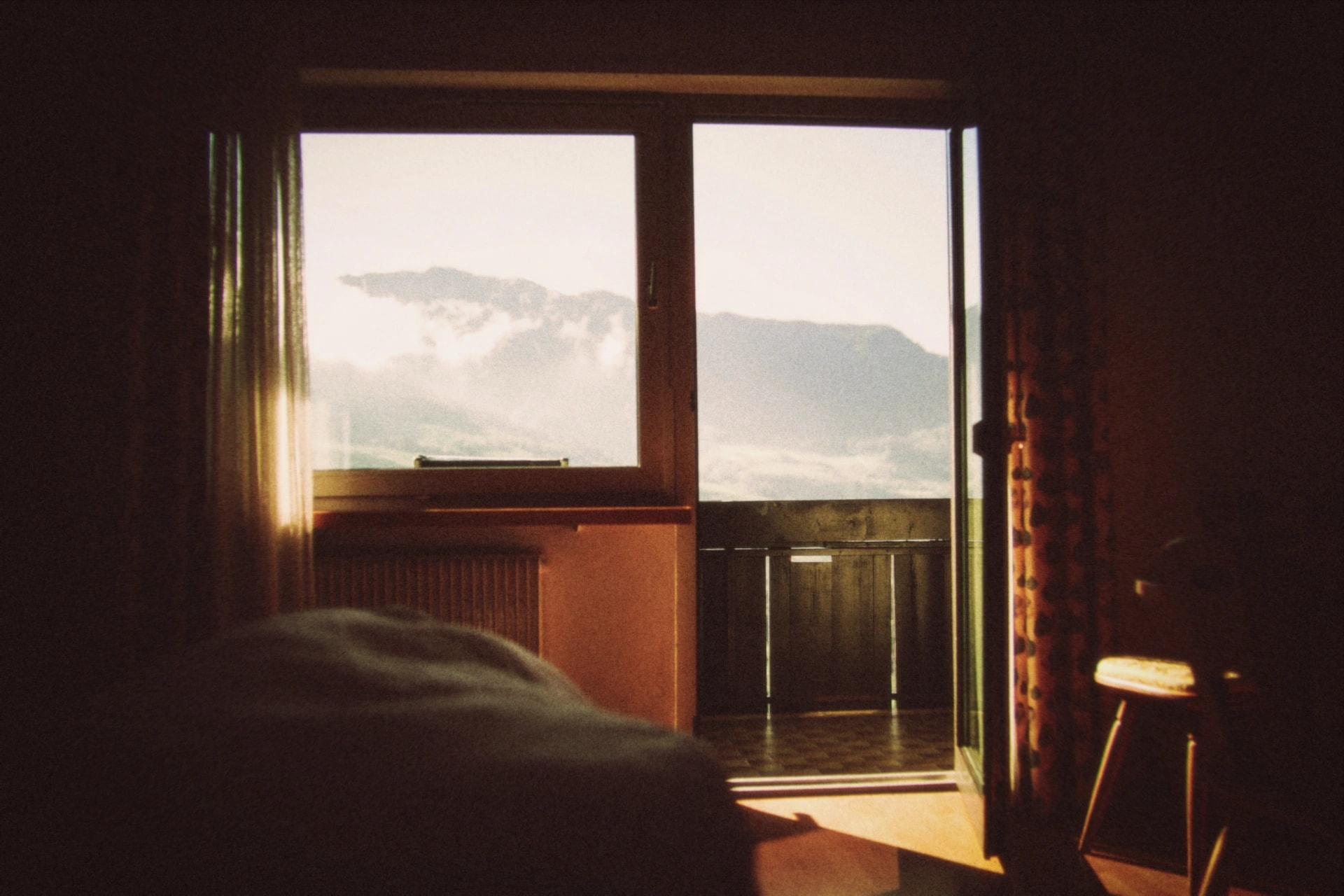A dim bedroom looking out through open balcony doors to clouds over the mountains, rendered with the Nostalgia preset