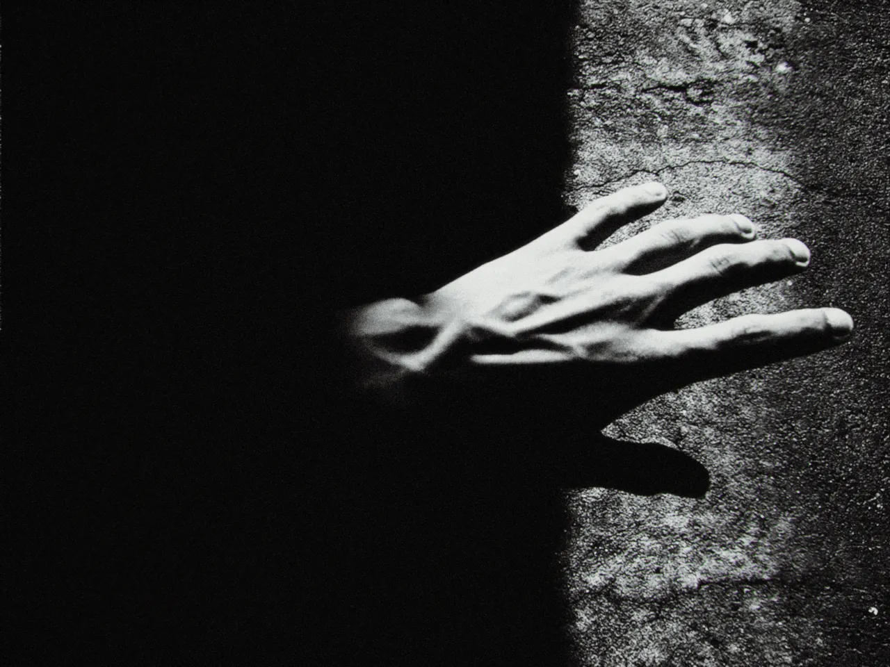 A hand pressed flat against a rough stone wall, half in shadow, in black and white, with contrast at 0