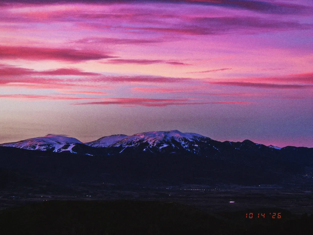 A pink and violet sunset over snow-capped mountains and a dark valley, date stamp on