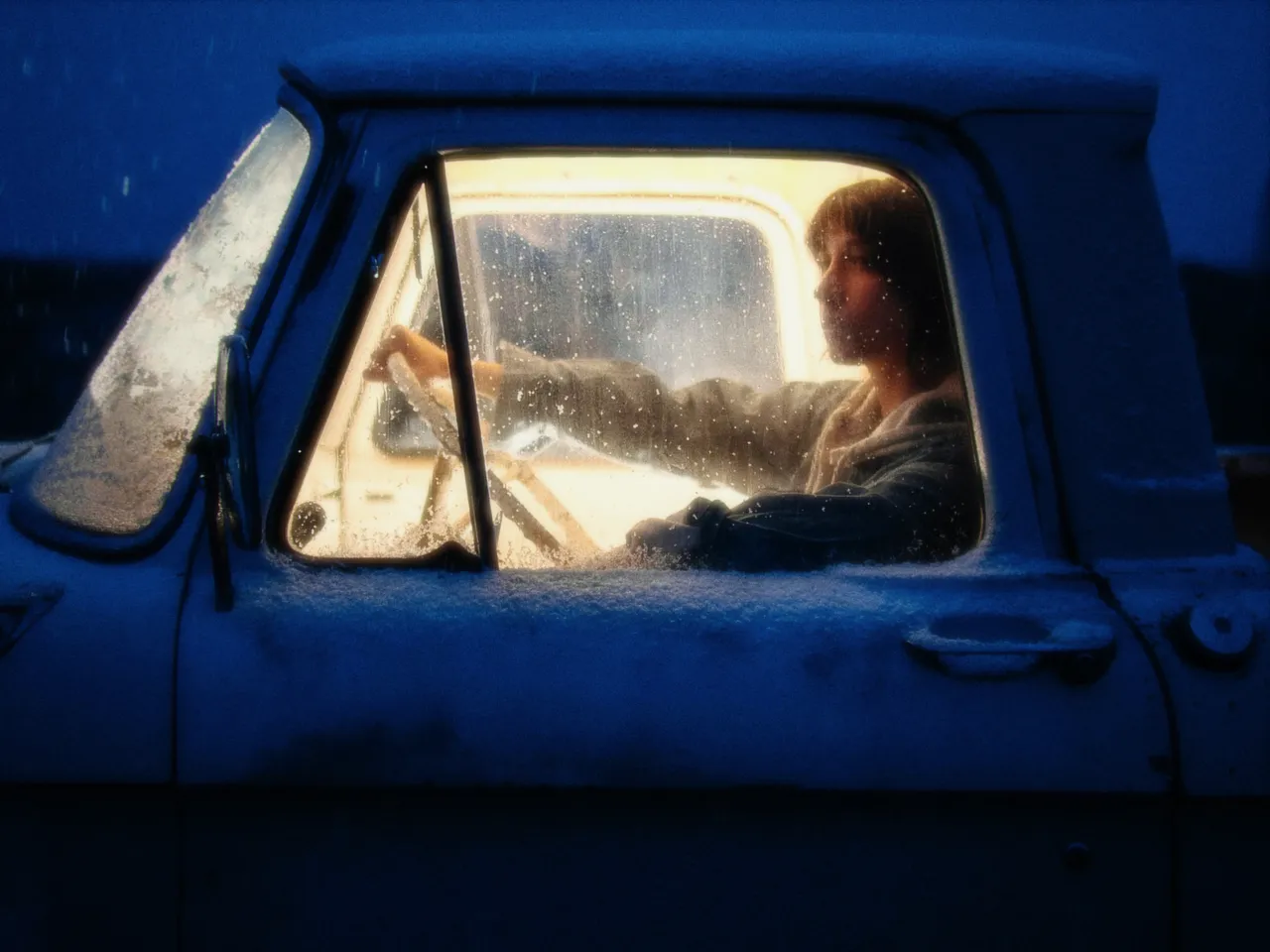 A woman at the wheel of an old blue truck at dusk, the cab lit warm from inside, with glow at 50%