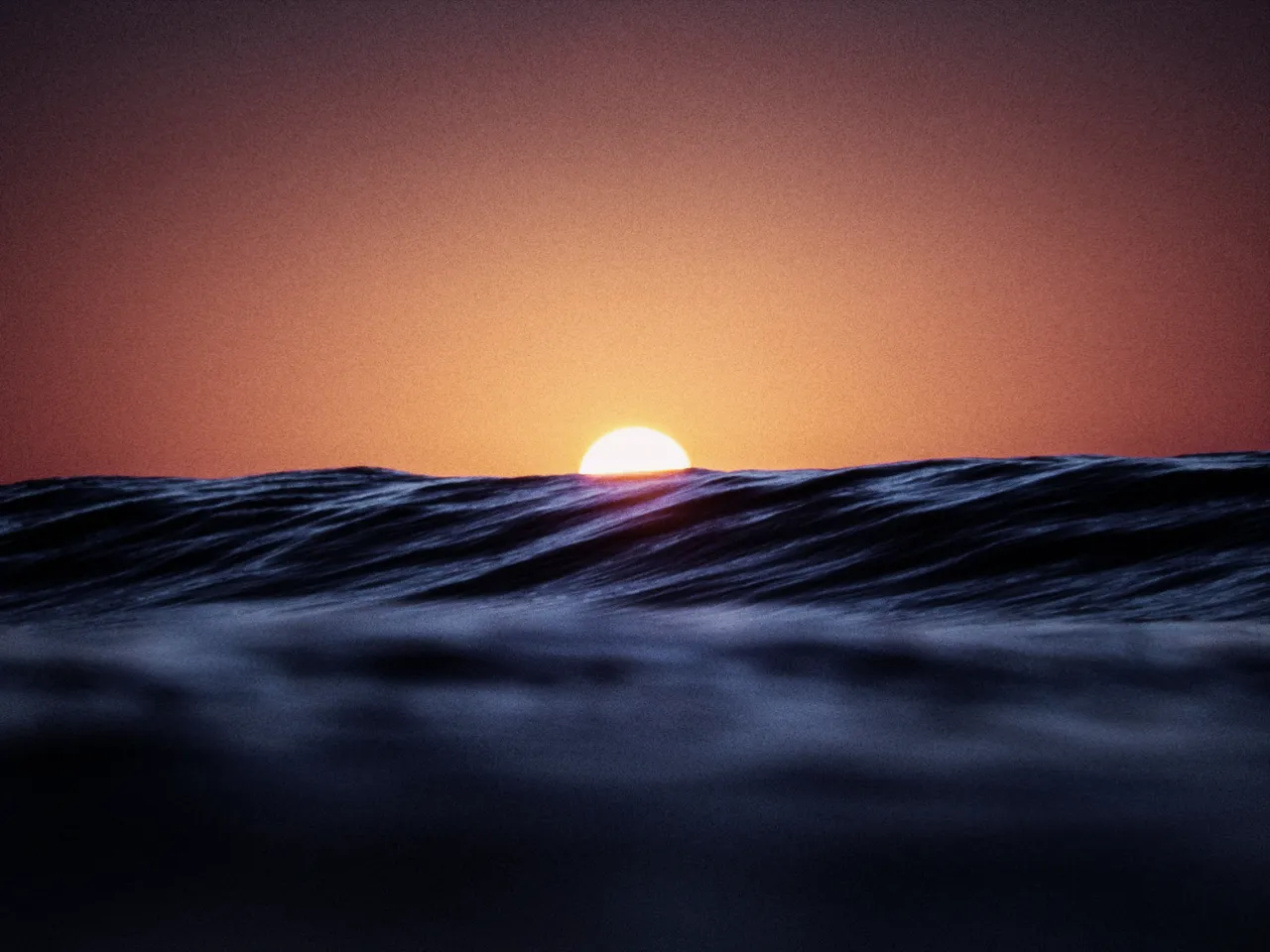 The sun sinking behind a dark ocean wave at dusk, with saturation at 0
