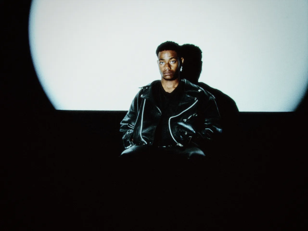 A man in a leather jacket seated in front of a huge glowing white circle, with vignette at 50%