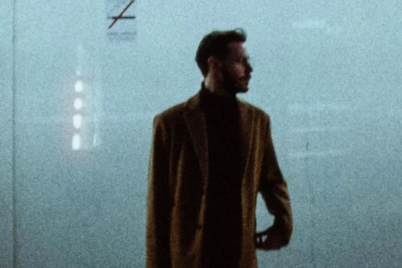 ASA 800 grain across the shelter wall, a close crop of a man in a long coat waiting under the strip lights of a bus shelter at night