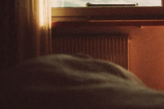 Film fog lifts the dark room into a warm haze, a close crop of a dim bedroom looking out through open balcony doors to clouds over the mountains
